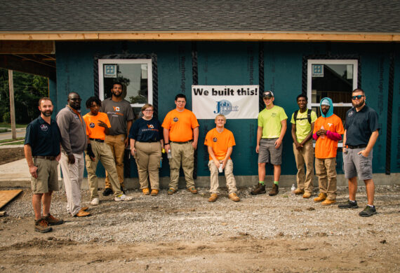 Students stand in front of a house under construction next to a sign that says "We built this"
