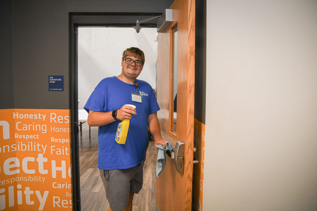 A young man stands in the doorway of a class room holding cleaning solution and a wash cloth. He is smiling.