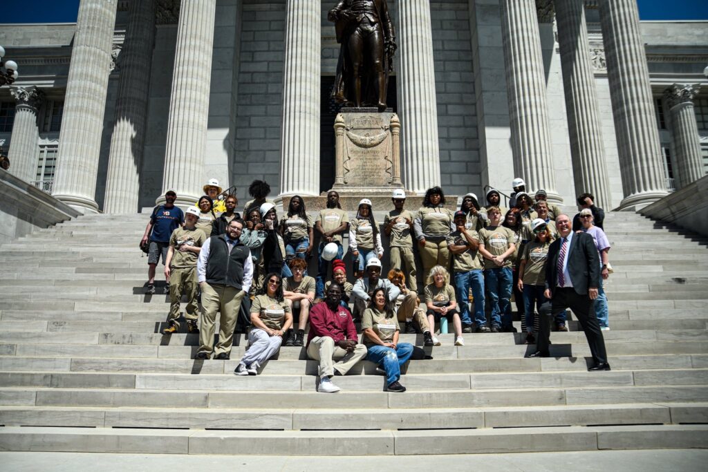 A group of people sit together on the steps of the Missouri State Capitol. The sun is shining and there are dark shadows on most faces.
