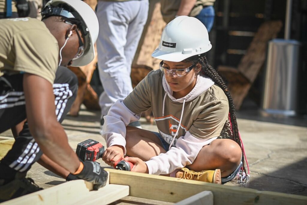 Ms. Hicks sits on the floor drilling a screw into the wall.