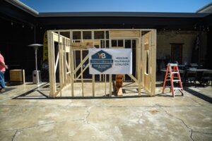 A bare framed room with four walls stands in an open outdoor space with a sign that says "Missouri YouthBuild Coalition."