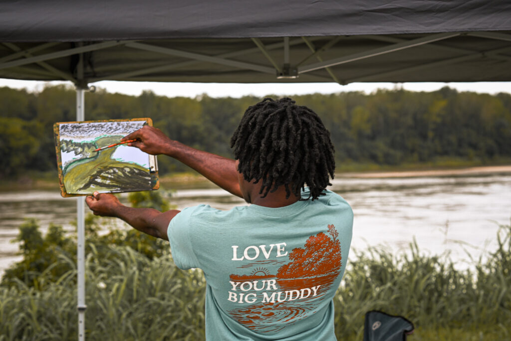 A young man holds up his painting and attempts to paint standing up